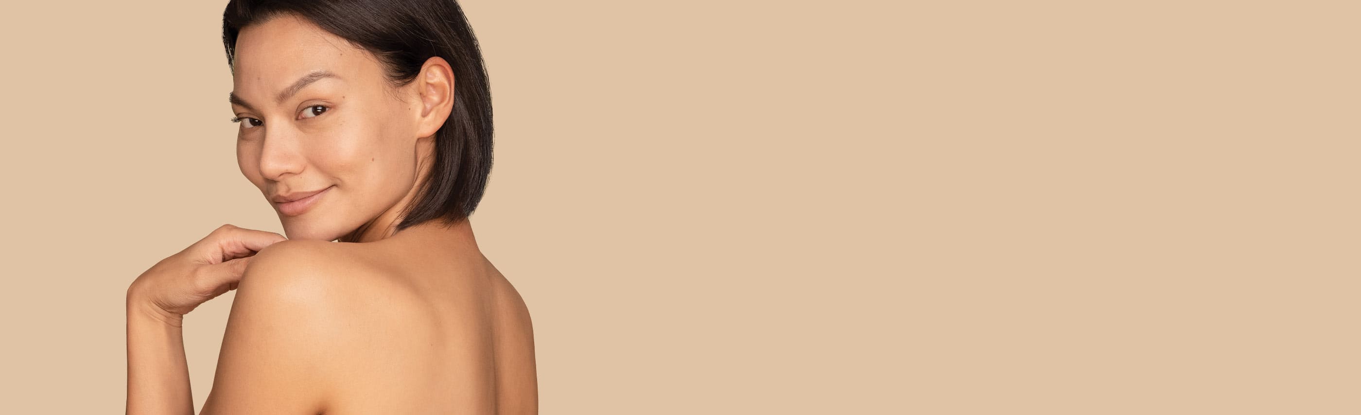 Femme aux cheveux noirs courts et au dos nu regardant par-dessus son épaule gauche en souriant.